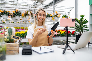 A florist uses a phone for a live sale of gardening tools in her shop, offering gardening advice...