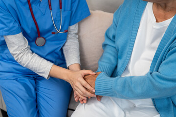 Close-up of young Asian nurse gently holding hand of elderly Asian patient on sofa. Nurse wearing...