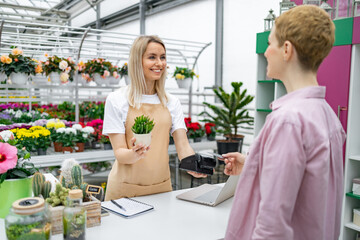 A florist helps a customer pay for a purchase at the checkout, with a smile, holding a small plant in a pot.