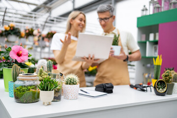 Two florists look at a laptop on a table in their shop, surrounded by various potted plants and cacti, managing their business.