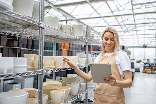 A shop assistant with a tablet inspects shelves stocked with various flowerpots, preparing for inventory.
