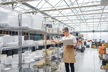 A Caucasian man inspects flowerpots in a shop, holding a laptop and an apron on. Rows of plant pots fill the shelves.