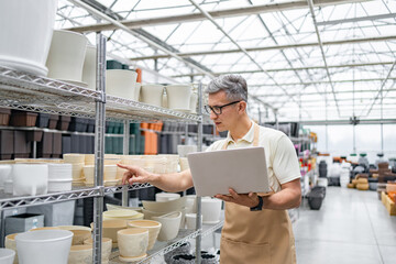 A man in a shop with a laptop looks at the flowerpots on the shelves, browsing and making a choice.
