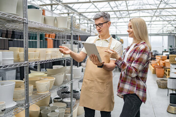 A Caucasian woman discusses flower pots with a shop assistant at a plant store. They're examining options on a tablet device near shelves.