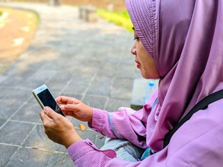 Muslim woman wearing pink hijab using smartphone while relaxing in outdoor park