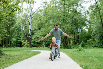 Toddler riding balance bike with dad cheering behind