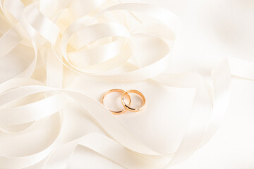 Two gold classic wedding rings lie among beautiful twisted beige ribbons on a satin background. A copy of the space. Chic wedding design layout.
