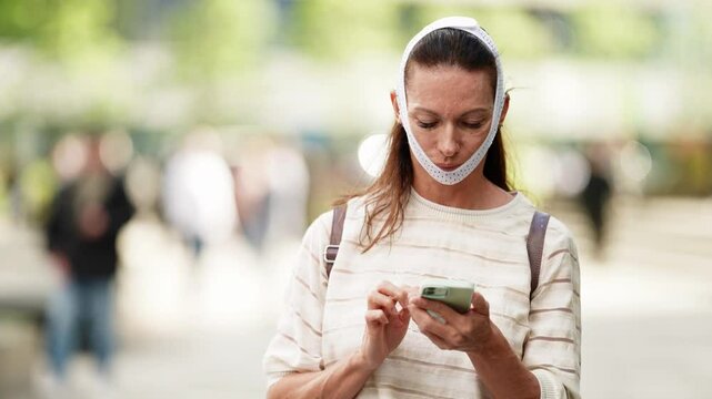 Woman wearing a white compression face band outdoors, looking down at her smartphone, blurred city background.