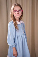 Backlit portrait of a young girl with long curly hair wearing glasses in a pink frame. She is wearing a blue dress decorated with buttons and a white lace collar. In the background is a light curtain.
