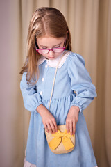 A young girl with long blonde hair is looking for something in her small straw bag with a yellow bow. The atmosphere of the photo conveys a sense of childish naivety, creating a warm mood.