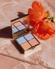 Beautiful eyeshadow palette featuring shades of blue, pink, and peach, elegantly placed on marble surface alongside vibrant orange flowers, creating stunning visual contrast