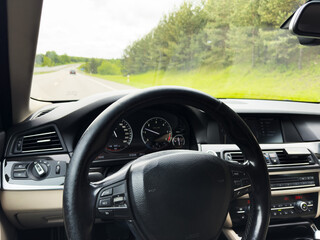 Front-seat perspective of a modern car interior, highlighting the steering wheel, digital dashboard and climate controls on an open highway.  Road trip by car.