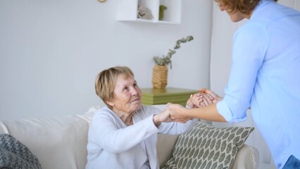 Young nurse assisting a senior woman in getting up from the sofa in the living room, providing compassionate support and care to enhance the elderly patient's comfort and independence at home