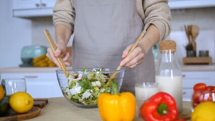 Female chef mixing a vibrant, fresh salad in a glass bowl using wooden spoons, surrounded by an array of colorful vegetables, fruits, and a bottle of milk in a cozy kitchen