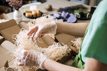 Gloved hands arrange green and lavender soaps with dried flowers in kraft packaging. Handmade...