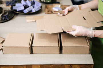 Person folds kraft boxes at a table near handmade soaps and eco filler. Scalable packaging, modular box system, ecommerce adaptability, minimal waste design