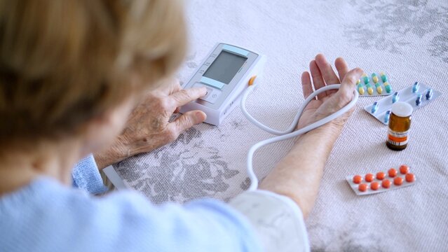 Senior woman using a digital blood pressure monitor at home, with various medications visible on the table beside her, emphasizing the importance of managing health in old age