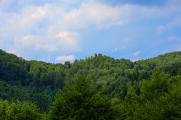 Rows of dense green forests cover the hills, creating a picturesque mountainous landscape under a bright blue sky with white clouds. On the horizon, through the treetops, silhouettes of ancient rocks 
