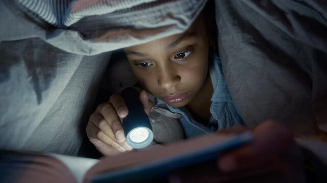 Zoom in shot of Black girl in pajamas hiding under blanket at night reading captivating book out loud using torch