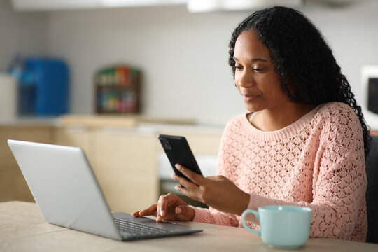 Serious black woman using cell phone and laptop in a kitchen
