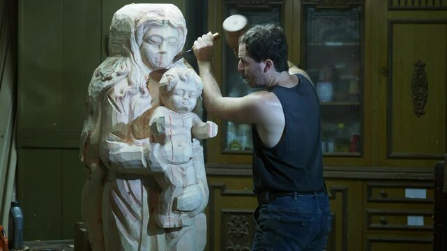 Skilled strong caucasian sculptor carving wooden virgin mary religious statue with mallet and chisel in workshop, showcasing creativity and craftsmanship in close up shot, slow motion