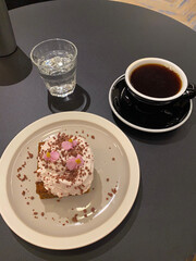 Minimalist black table setup in a specialty coffee shop with black coffee, dessert with cream and edible flowers, and glass of water – modern café scene