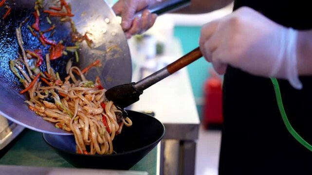 Chef plating stir-fried noodles with vegetables and chicken into a black bowl, ready for serving.