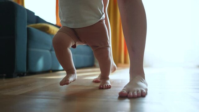 Mother supports son taking first step. Baby boy barefoot learning to walk. Mother hands guide son first steps. Son walks barefoot for first time. Tender family moment. Mother helping son take step.