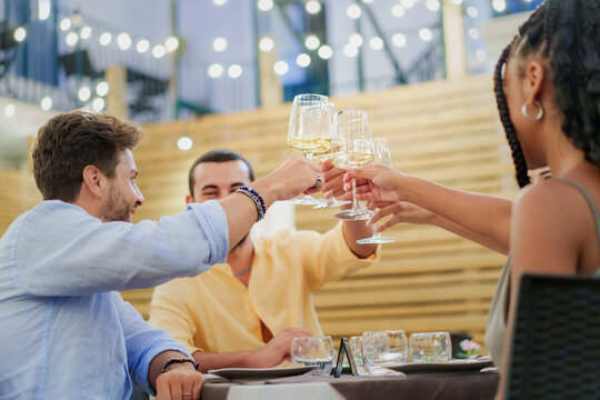 Group of diverse friends toasting with white wine at a restaurant party at night with bokeh lights.