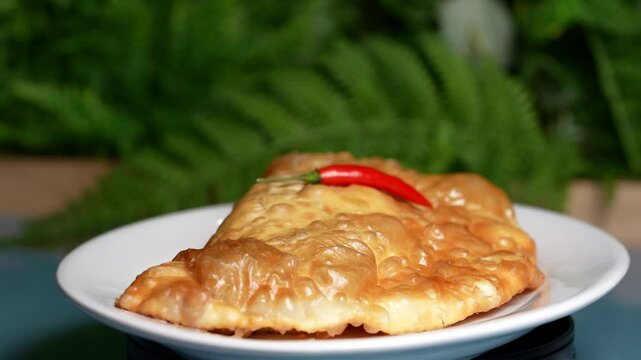 Crispy cheburek with puffy golden crust and bright red chili pepper on top, against a green leafy background.