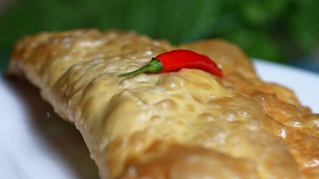 Golden crispy cheburek pastry topped with a single red chili pepper, served on a white plate.