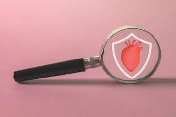 A magnifying glass examining a protected heart symbol on pink background.,Represents health insurance, life insurance,heart health, cardiology, cardiovascular check-up,healthcare and medical.