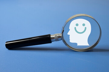 Magnifying glass focusing on a human head icon with a smiling face on blue background. Concept for mental health awareness, psychological well-being, positive thinking, emotional health.