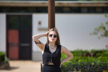 Young, beautiful blonde woman with tattoos, sunglasses and dressed in black leaning against a...