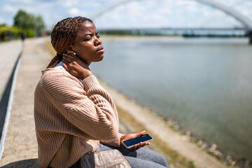 Young beautiful black woman is sitting by the river in the city. She has pain in her neck.