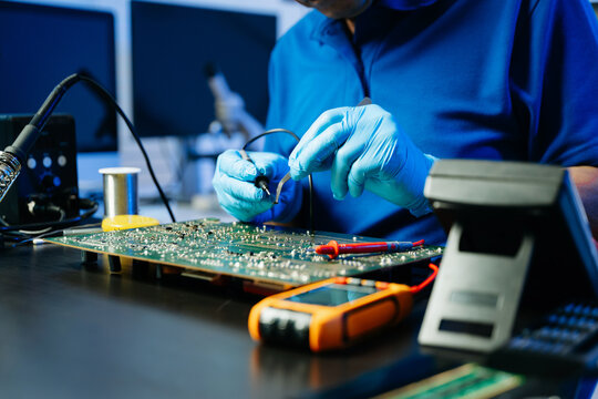 Electronics technician soldering a green PCB with precision in a modern lab, wearing gloves and surrounded by digital tools