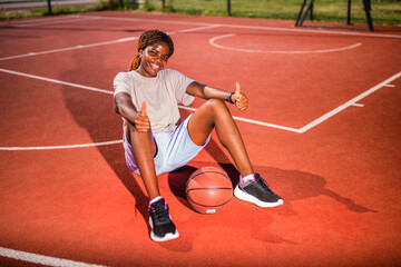 Obraz premium Young black woman practicing basketball alone at sports court in the morning. Happy girl sitting on basketball court on sunny day.