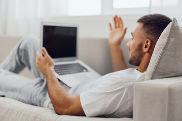 Young man relaxing on a couch, engaging in a video call while working on a laptop. Modern, cozy home interior with bright light and soft furnishings.