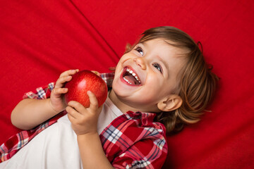Adorable kid holding apple fruit and smiling over red studio background. Healthy relationship with food and child nutrition concept. Healthy baby food with vitamins and kids vegetarianism. 