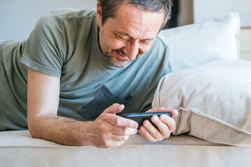 Casual male watching entertaining social media content on mobile phone device while laying on living room sofa