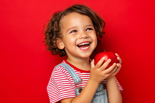 Adorable kid holding apple fruit and smiling over red studio background. Healthy relationship with food and child nutrition concept. Healthy baby food with vitamins and kids vegetarianism. 