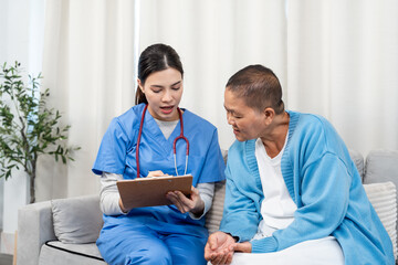 Obraz premium Young Asian nurse wearing blue uniform discussing medical notes on clipboard with elderly Asian man on sofa. Nurse explaining with hand gesture while patient listening attentively. Home healthcare