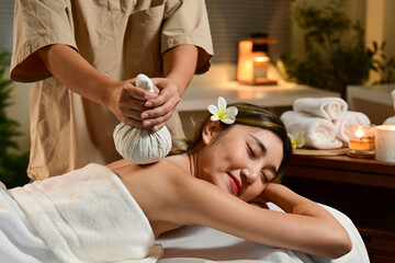 Lady receiving hot herbal ball spa massage, masseur gently compresses herb bags on woman back in warm lighting of candles at spa salon