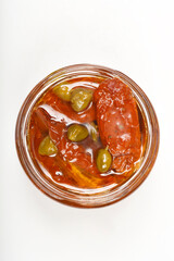 An overhead close-up shows sun-dried tomatoes and green capers preserved in oil within a glass jar, set against a plain background
