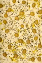 A close-up, top-down view shows raw focaccia dough topped with green olives, rosemary, black salt, and olive oil, ready for baking