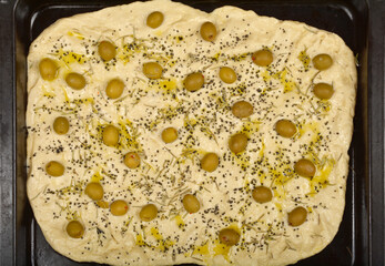 An overhead shot shows raw focaccia dough on a baking tray, topped with green olives, rosemary, black salt, and a drizzle of olive oil, ready for baking