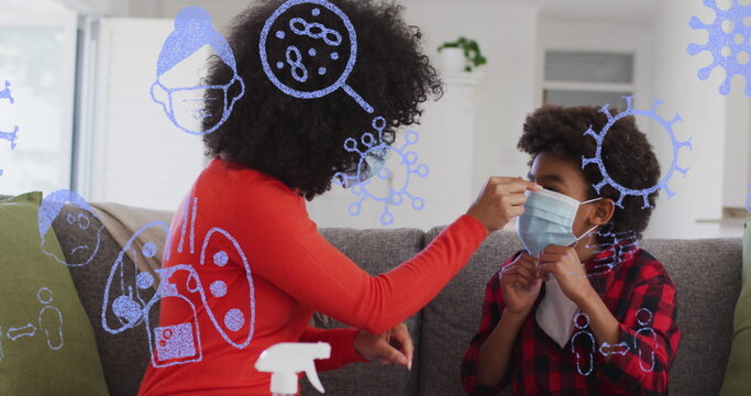 Image of pandemic icons over african american mother and son in face masks sitting on sofa - Powered by Adobe