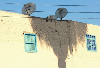 Wall of an old house with palm trees shadow and satellite dishes on rooftop.