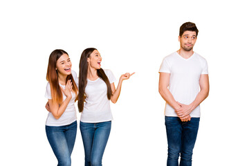 Fototapeta premium Portrait of excited girls mocking at guy standing shy wearing white t-shirt denim jeans isolated over yellow background