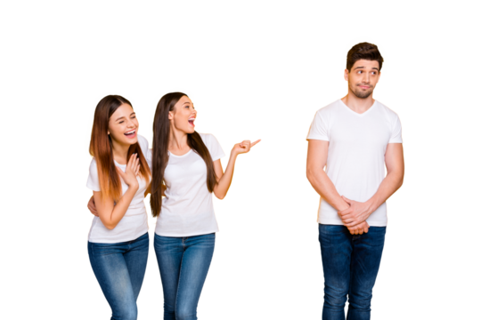 Portrait of excited girls mocking at guy standing shy wearing white t-shirt denim jeans isolated over yellow background